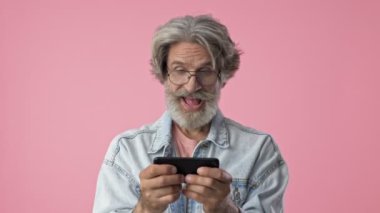Excited elderly stylish bearded man with gray hair in denim jacket smiling and playing game on smartphone over pink background isolated