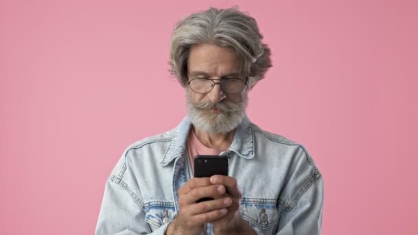 デニムジャケットに白髪の驚きのおしゃれなあごひげの男が ピンクの背景の上にスマートフォンを使用しながら幸せになり 笑顔になる — ストック動画
