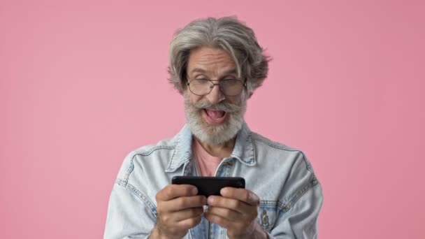 デニムジャケットで白髪の興奮したお年寄りスタイリッシュなあごひげの男が笑顔で ピンクの背景の上にスマートフォンでゲームをプレイ孤立 — ストック動画