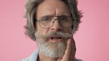 Pembe arka plan izole üzerinde kameraya bakarken diş ağrısı nedeniyle eliyle çenesini dokunan denim ceket gri saçlı üzgün yaşlı şık sakallı adam yakından görünümü