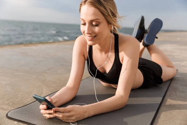 Imagen de una mujer sonriente escuchando música en el teléfono celular mientras está acostada en la alfombra cerca de la playa por la mañana — Foto de Stock