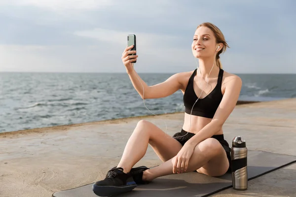 Kép pozitív nő segítségével a fülhallgató és a mobiltelefon mellett ül szőnyeg közelében tengerparton reggel — Stock Fotó