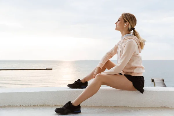 Image of attractive woman listening to music with earphones while sitting near seaside in morning — Stock Photo, Image