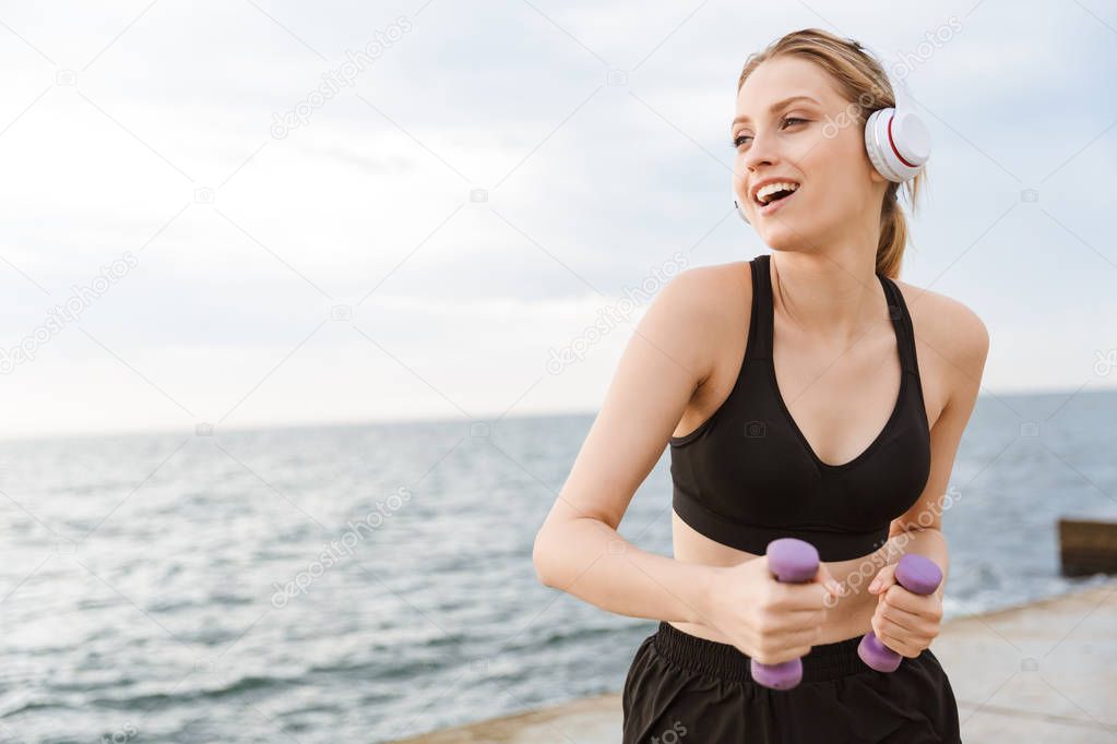 Image of smiling cute woman using headphones while working out with dumbbells on pier near seaside in morning