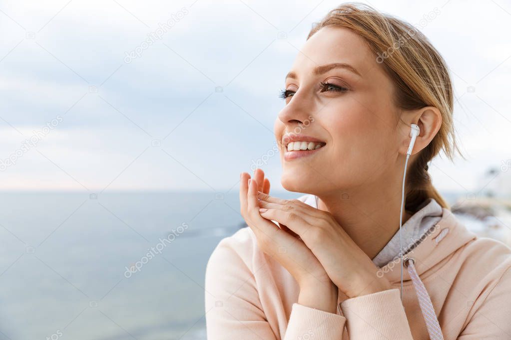 Image of nice woman smiling and listening to music with earphones while sitting near seaside in morning