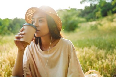 Yeşil parkta çimenüzerinde paket kahve içerken kulaklık kullanan kuluçkaya yakan kadının portresi