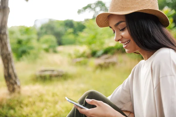 Porträt einer charmanten Frau mit Strohhut, die lächelt, während sie ihr Smartphone im grünen Park hält — Stockfoto