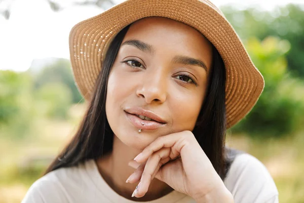 Foto de una mujer hermosa con sombrero de paja mirando a la cámara mientras camina en el parque verde — Foto de Stock