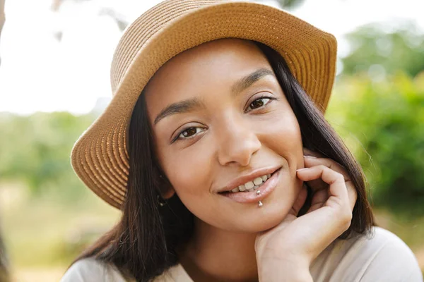 Foto de mujer alegre con sombrero de paja sonriendo a la cámara mientras camina en el parque verde —  Fotos de Stock