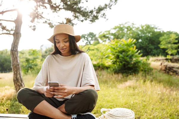 Портрет привабливої жінки в солом'яному капелюсі, що тримає смартфон, сидячи на лавці в зеленому парку — стокове фото