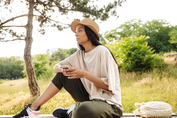 Portré gondolkodó nő visel Szalmakalapot gazdaság okostelefon, miközben ül a padon zöld parkban — Stock Fotó
