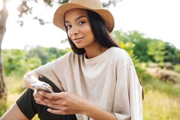 Retrato de mulher bonita usando chapéu de palha segurando smartphone enquanto sentado no banco no parque verde — Fotografia de Stock