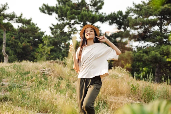 Φωτογραφία της νεαρής γυναίκας που δείχνει το δάχτυλο στο ακουστικό και ακούγοντας μουσική στο κινητό τηλέφωνο ενώ περπατάει στο πράσινο πάρκο — Φωτογραφία Αρχείου