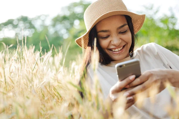 Retrato de mulher engraçada usando chapéu de palha digitando no celular enquanto estava deitado na grama no parque verde — Fotografia de Stock