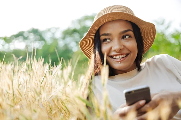 Retrato de mulher feliz usando chapéu de palha digitando no celular enquanto deitado na grama no parque verde — Fotografia de Stock