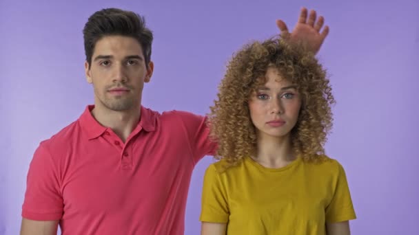 ハンサムな若いブルネットの男は 孤立した紫色の背景の上にカメラを見ながら 彼女の頭の上に手で彼の若いかわいいガールフレンドを置きます — ストック動画