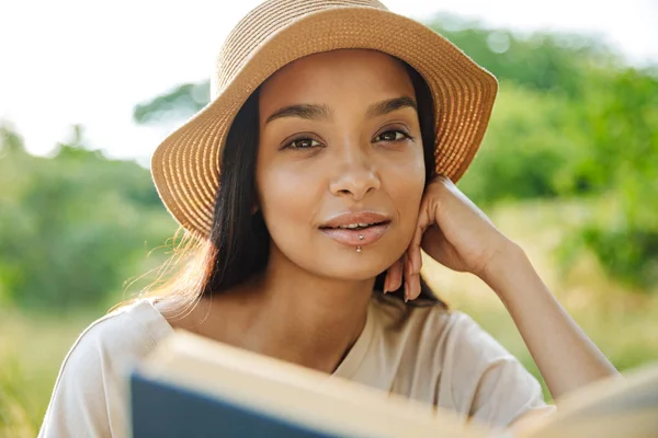 Portrét atraktivní ženy v slaměném klobouku čtení knihy při sezení na trávě v zeleném parku — Stock fotografie