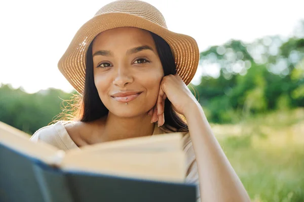 Porträt einer lächelnden Frau mit Strohhut, die Buch liest, während sie im grünen Park auf Gras sitzt — Stockfoto