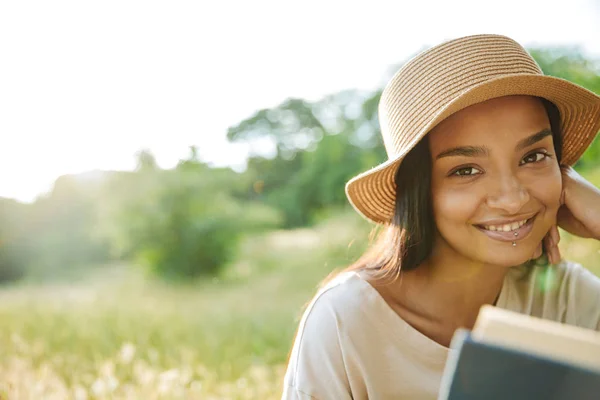 Porträt einer fröhlichen Frau mit Strohhut, die Buch liest, während sie im grünen Park auf Gras sitzt — Stockfoto