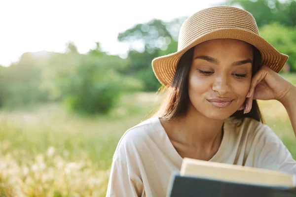 Porträt einer zufriedenen Frau mit Strohhut, die Buch liest, während sie im grünen Park auf Gras sitzt — Stockfoto
