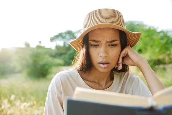 Porträt einer ratlosen Frau mit Strohhut, die im grünen Park auf dem Gras sitzt und Buch liest — Stockfoto