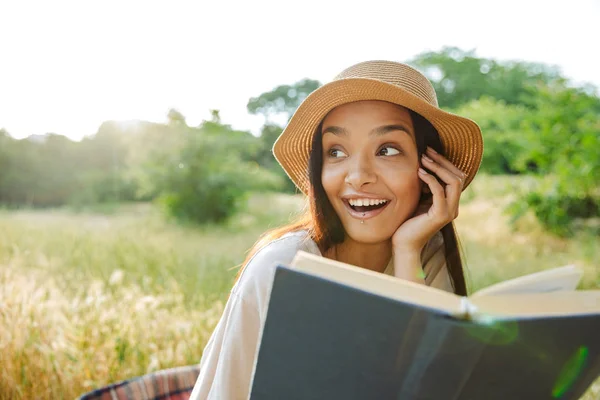 Portrait of delighted woman wearing straw hat reading book while sitting on grass in green park — Stock Photo, Image