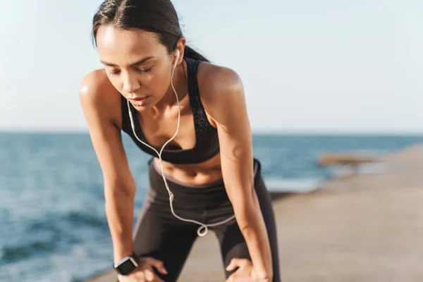 Serio hermoso joven fuerte deportivo mujer al aire libre en la playa por la mañana tener un descanso escuchando música con auriculares . —  Fotos de Stock