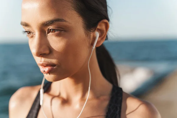 Increíble mujer deportiva fuerte concentrada al aire libre en la playa por la mañana posando escuchando música con auriculares . — Foto de Stock