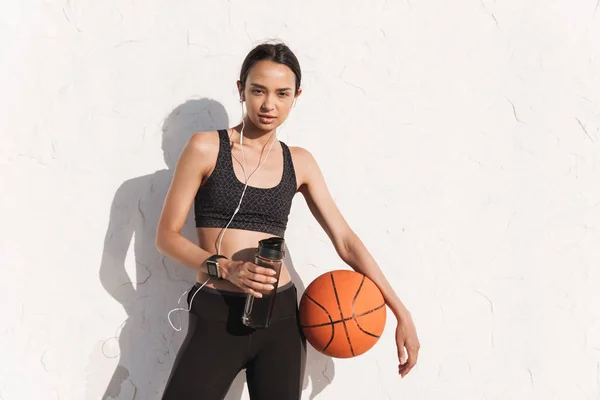 Incrível concentrado forte esportes mulher ao ar livre sobre branco parede fundo posando ouvir música com fones de ouvido segurando bola e garrafa com água . — Fotografia de Stock