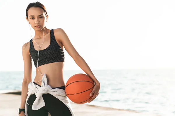 Bela mulher forte esportes ao ar livre na praia posando ouvir música com fones de ouvido segurando basquete . — Fotografia de Stock