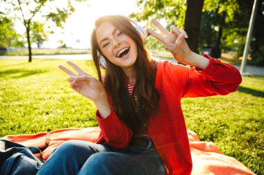 Yeşil parkta otururken kulaklık ve gesturing barış işareti ile müzik dinleneşeli kız öğrenci görüntü