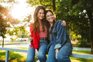 Yeşil parkta korkuluk üzerinde otururken gülümseyen ve sarılan iki mutlu kız Görüntü