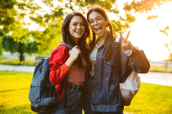 Image of two laughing girls students looking at camera and gesturing peace sign while walking in green park — Stock Photo, Image