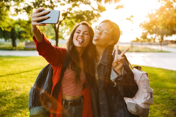 Imagen de dos chicas divertidas con lenguas colgando tomando foto selfie en el teléfono celular y haciendo gestos de señal de paz mientras camina —  Fotos de Stock