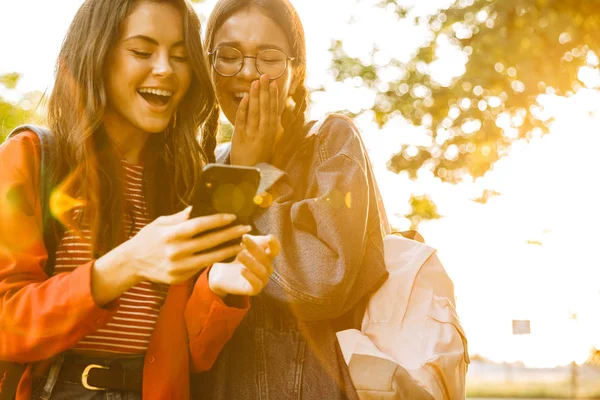 Immagine di due ragazze deliziate che ridono e scrivono sul cellulare mentre camminano nel parco verde — Foto Stock