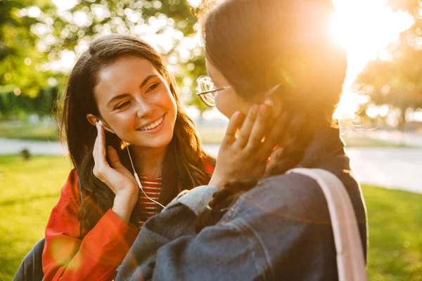 Image of two cute girls students using earphones and looking at each other while walking in green park — Stock Photo, Image