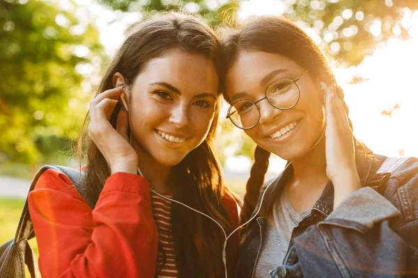 Image closeup of two cute girls students using earphones and looking at camera while walking in green park — Stockfoto