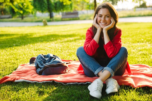 Imagen de una buena estudiante sonriendo y apoyando su cabeza mientras está sentada sobre una manta en el parque verde —  Fotos de Stock
