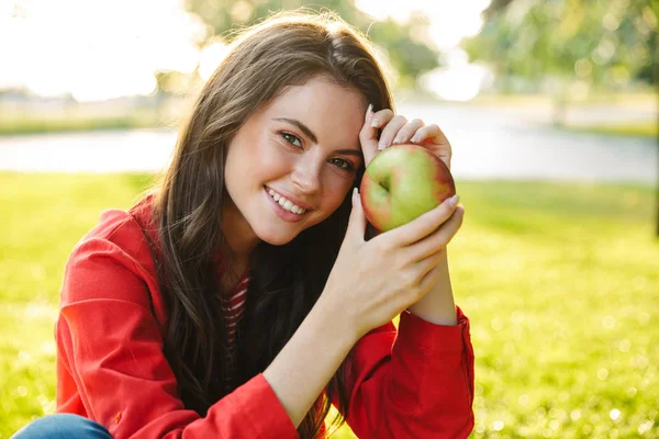 Image of nice girl student smiling and holding apple while sitting in green park — Stock Photo, Image