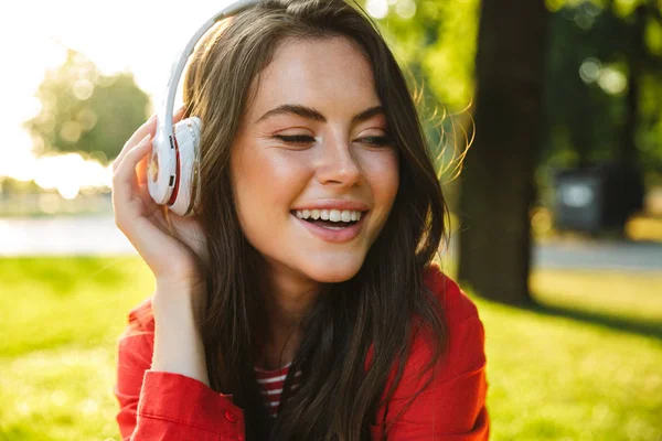 Imagen de primer plano de una estudiante sonriente escuchando música con auriculares mientras está sentada en el parque verde —  Fotos de Stock