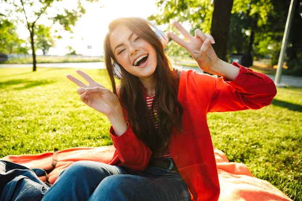 Imagen de una estudiante alegre escuchando música con auriculares y haciendo gestos de paz mientras está sentada en el parque verde —  Fotos de Stock