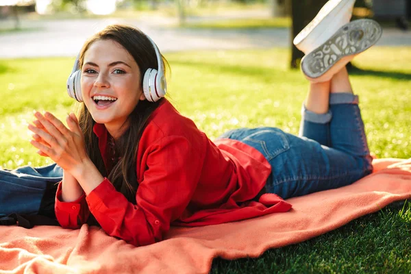 Image of delighted girl student listening music with headphones while lying on blanket in green park — Stock Photo, Image