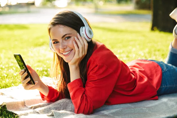 快乐的女学生用耳机听音乐，使用手机，而躺在绿色公园的毯子上的图像 — 图库照片