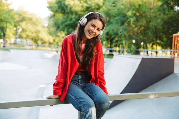 Spor sahasında korkulukta otururken kulaklıkla müzik dinleyen neşeli kız öğrencinin görüntüsü — Stok fotoğraf