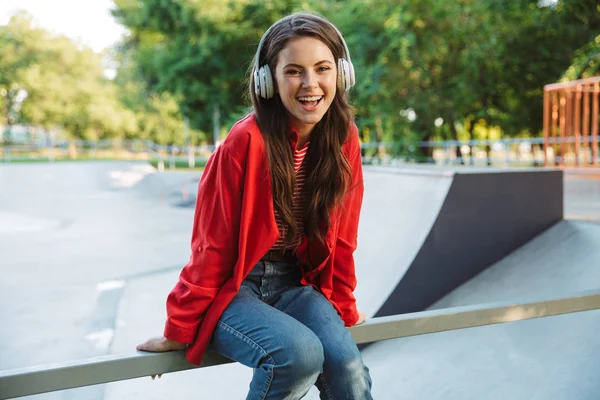 Imagen de estudiante alegre escuchando música con auriculares mientras está sentado en la barandilla en el campo de deportes —  Fotos de Stock