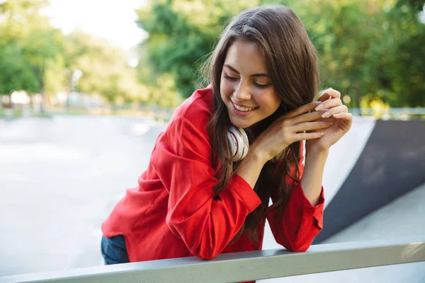 Kép Vértes az elégedett lány hallgató fejhallgató mosolyogva és hajlító a korlát a sportpálya — Stock Fotó