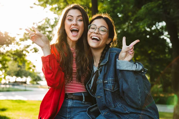 Image of two laughing girls gesturing peace sing and hugging while sitting on railing in green park — Stock Photo, Image