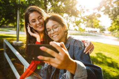 Yeşil parkta korkuluk üzerinde otururken cep telefonu selfie fotoğraf çekmek ve gülümseyen iki güzel kız görüntü