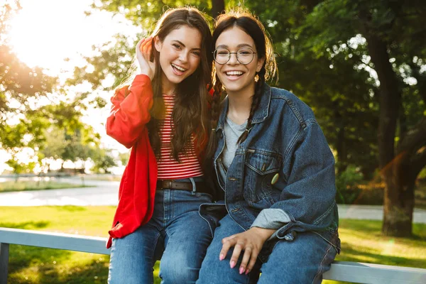 Immagine di due ragazze allegre che sorridono e si abbracciano mentre siedono sulla ringhiera nel parco verde — Foto Stock