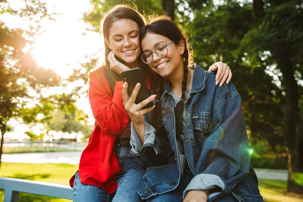 Εικόνα δύο χαμογελαστών κοριτσιών που χρησιμοποιούν κινητό και αγκάλιασμα ενώ κάθονται στο κιγκλίδωμα στο καταπράσινο πάρκο — Φωτογραφία Αρχείου